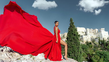 Woman in a striking red flying dress posing barefoot on a rocky hill with the Acropolis of Athens in the background, her dress billowing dramatically in the wind under a clear blue sky.