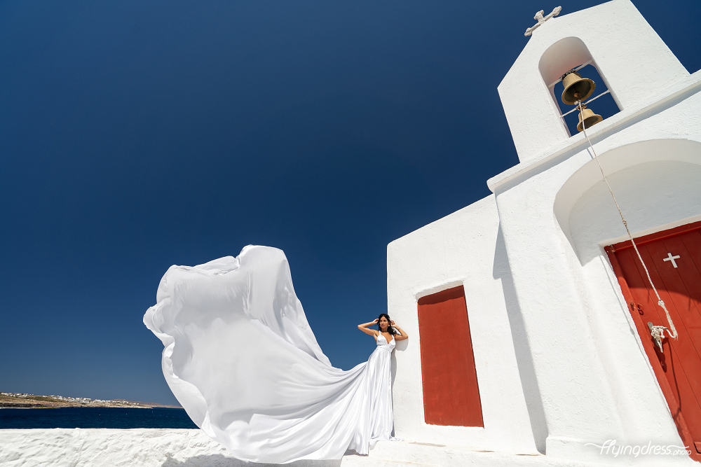 A woman in a flowing white dress poses dramatically in front of a white building with red doors and a bell tower against a clear blue sky in Mykonos.