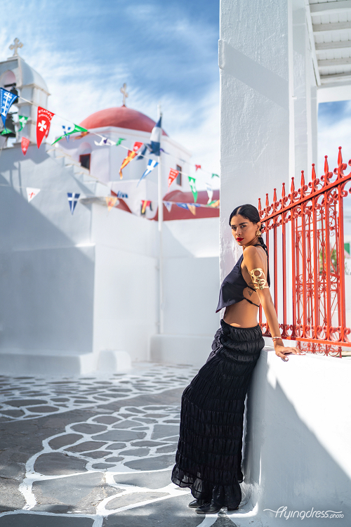 A woman in a chic black outfit leans casually against a white wall with red accents, set against the backdrop of a white-washed church adorned with colorful flags and a red dome in Mykonos, under a clear blue sky.
