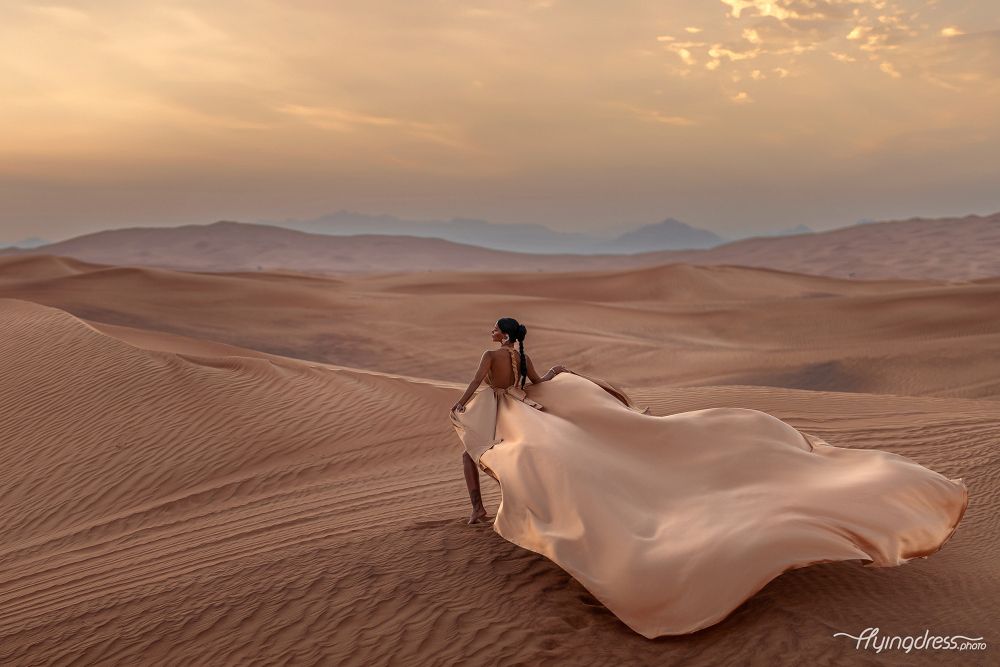 As the sun dips below the horizon, a model graces the Dubai desert in a flying dress photoshoot, capturing the enchanting blend of elegance and twilight hues against the golden sands.