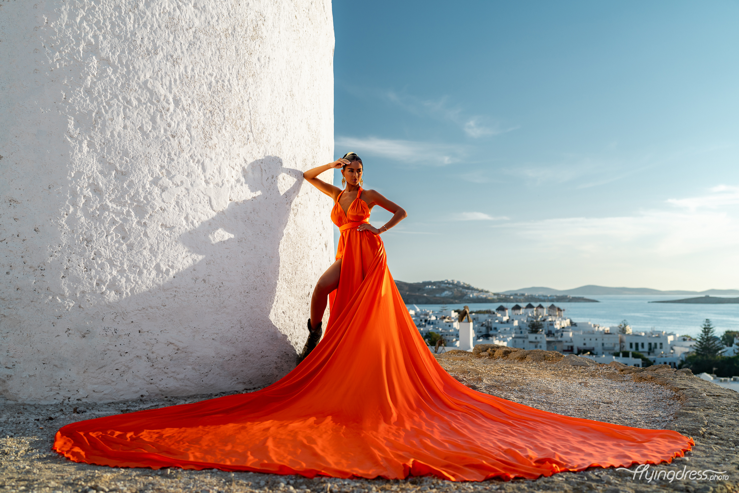 A woman in a striking orange gown poses confidently against a white-washed building in Mykonos, her dress flowing gracefully around her, with the beautiful Aegean Sea and white-washed buildings of the island in the distance.