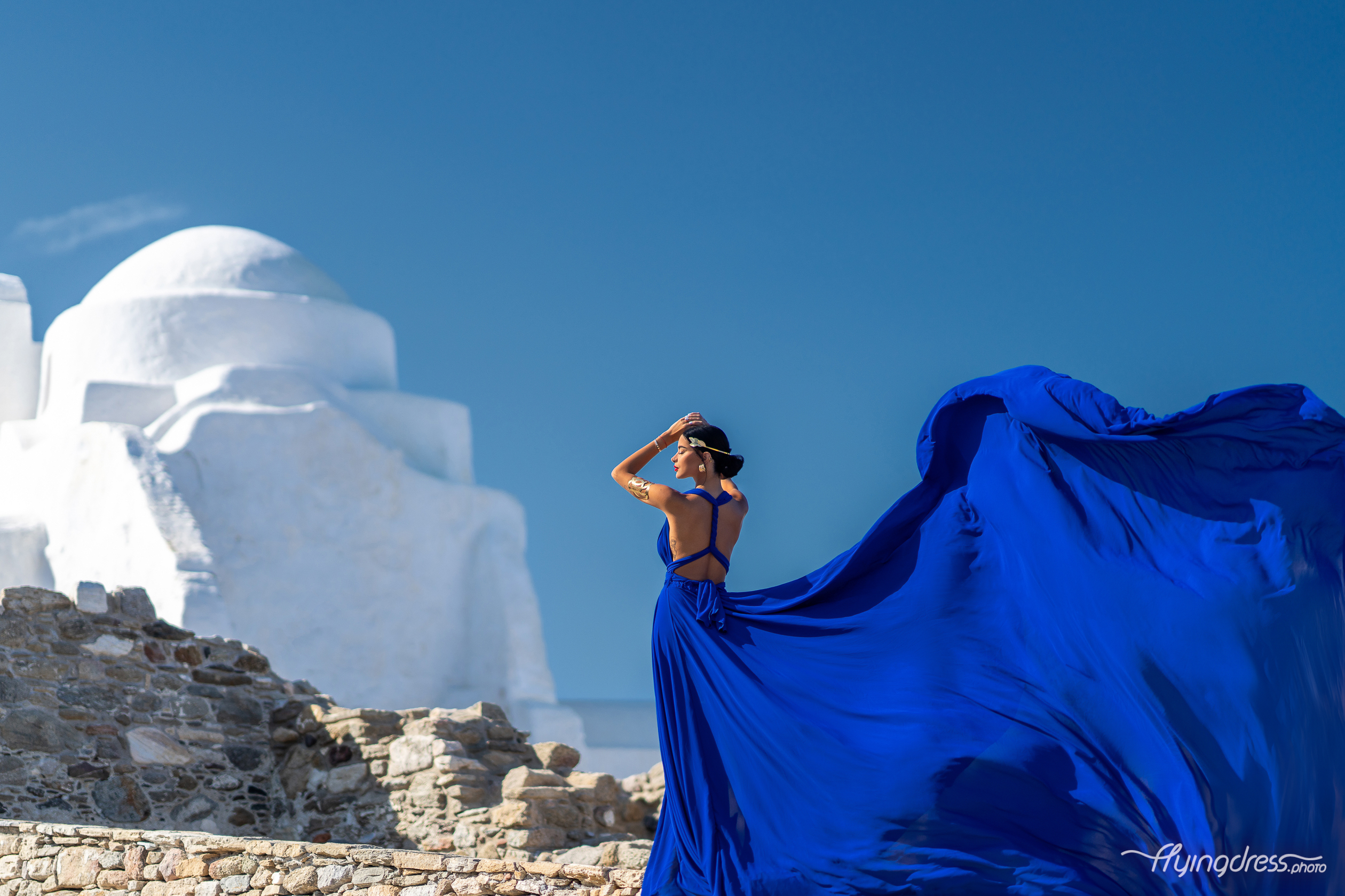 Woman in a royal blue flying dress showcasing the open back design, with the dress flowing dramatically in the wind against white-washed Mykonos architecture and a deep blue sky.