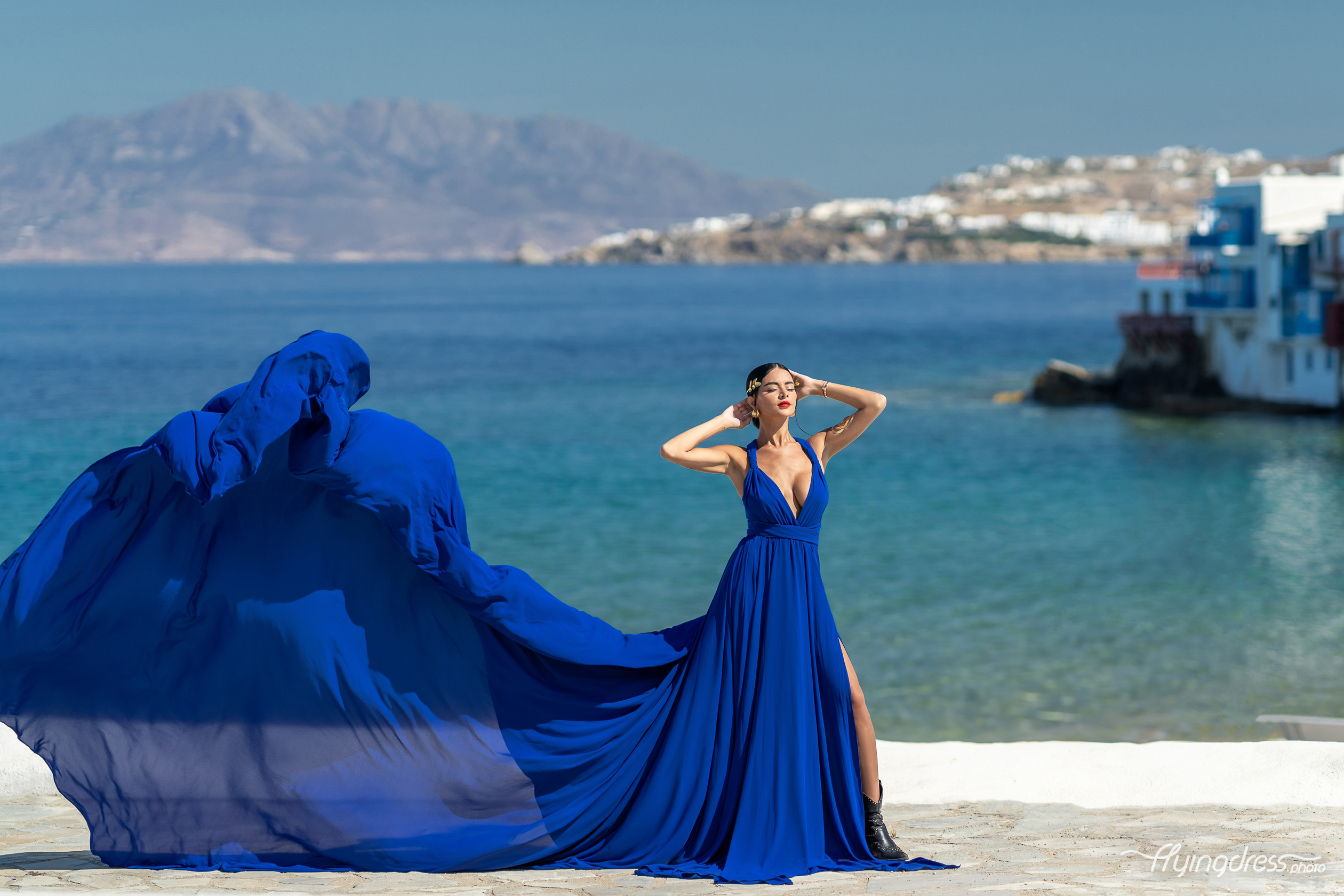Woman in a royal blue flying dress with a deep v-neckline posing in Little Venice, Mykonos, with the dress billowing dramatically against the turquoise sea and island backdrop.