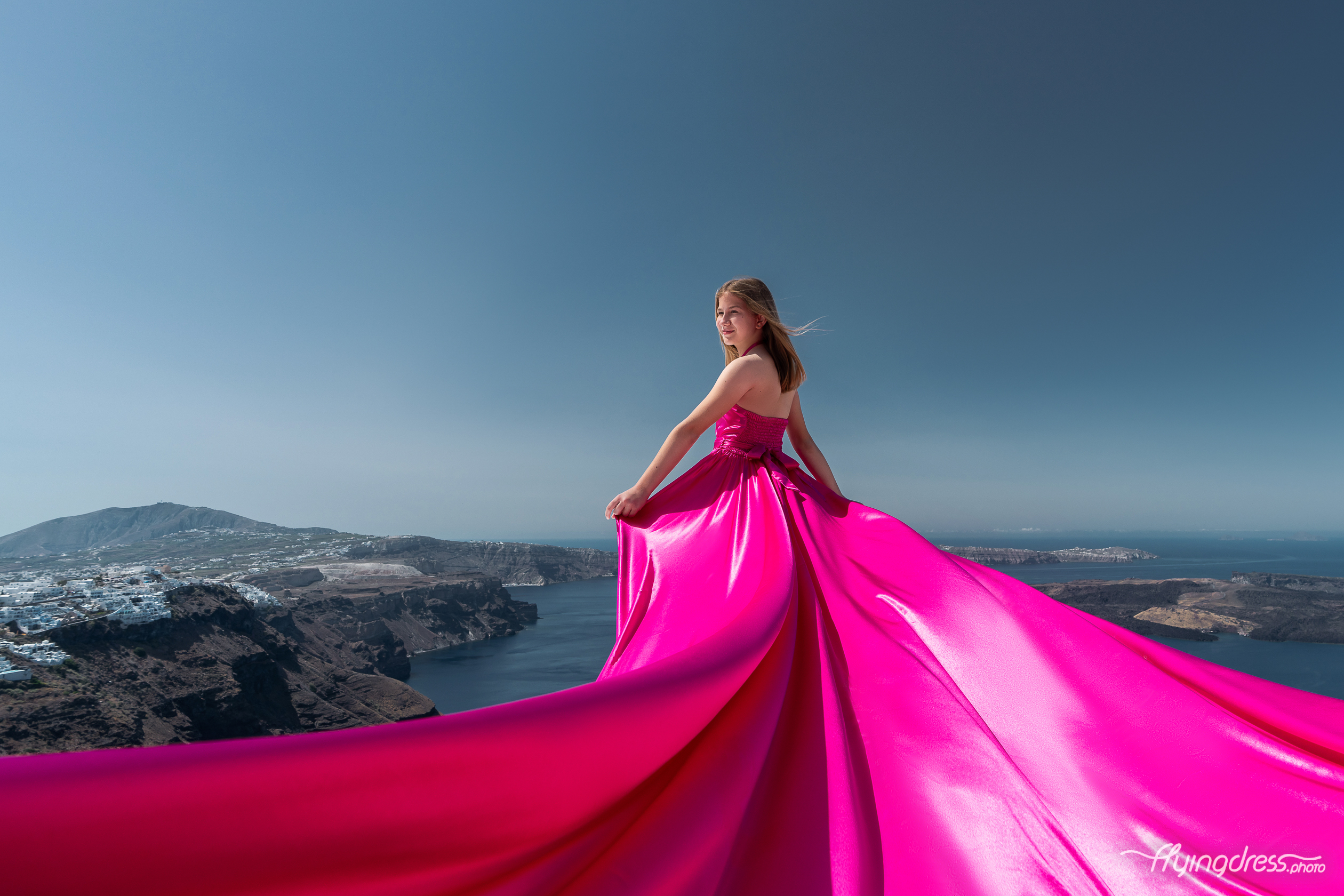 A girl in a stunning pink dress stands with the flowing train of her gown cascading behind her, set against the breathtaking coastal landscape of Santorini, Greece.