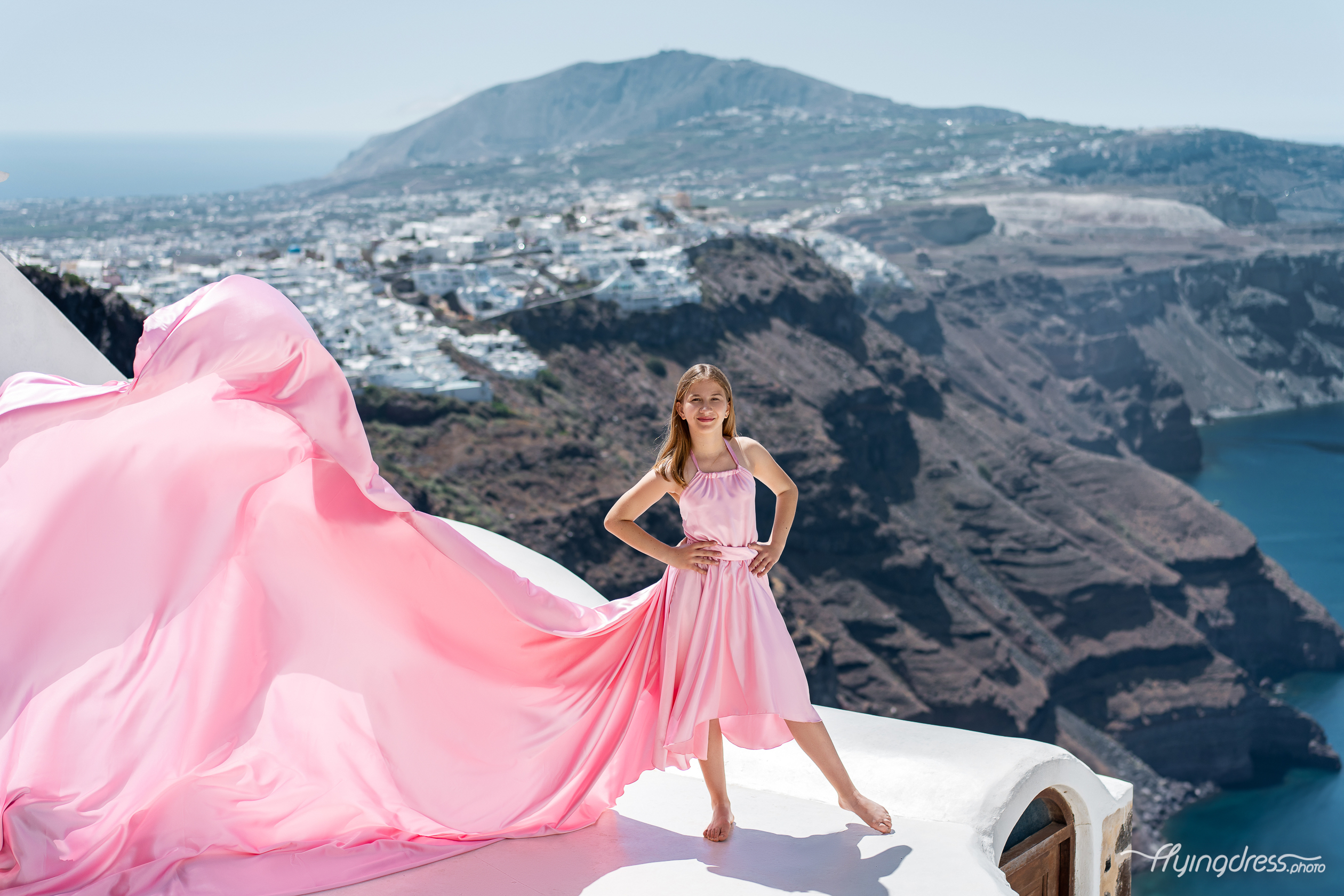 Make a statement with a gorgeous pink flying dress, showcasing a dramatic flowing train, perfect for capturing stunning photos against the breathtaking landscape of Santorini, Greece.