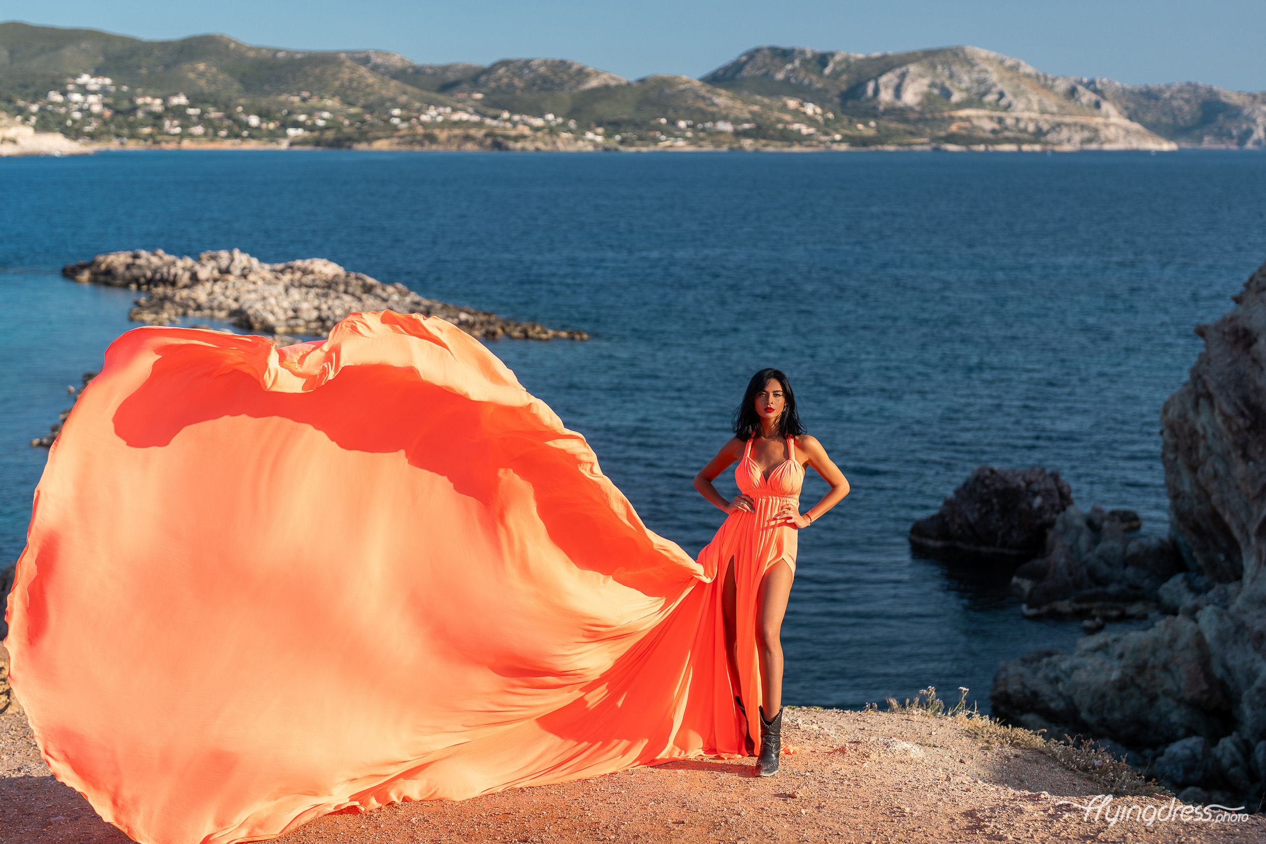 A woman in a bright orange flowing dress poses confidently on a rocky coastline, with the dress billowing dramatically against the backdrop of blue waters and distant hills.