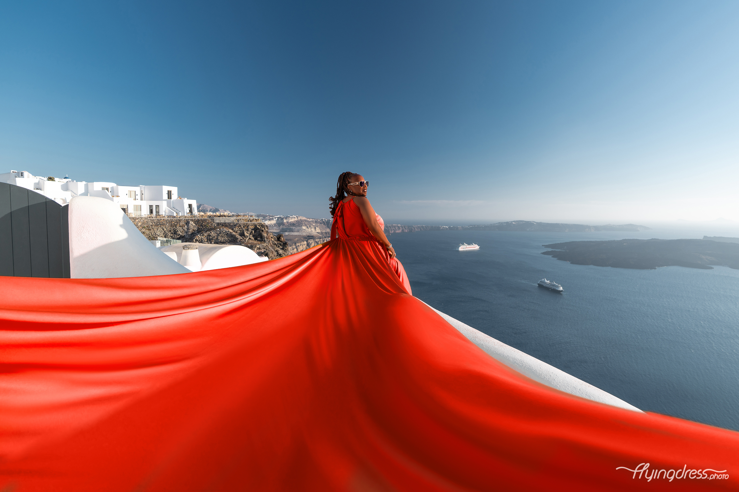 A scarlet flying dress sweeps dramatically over Santorini's white rooftops with the caldera and Aegean Sea stretching into the horizon.