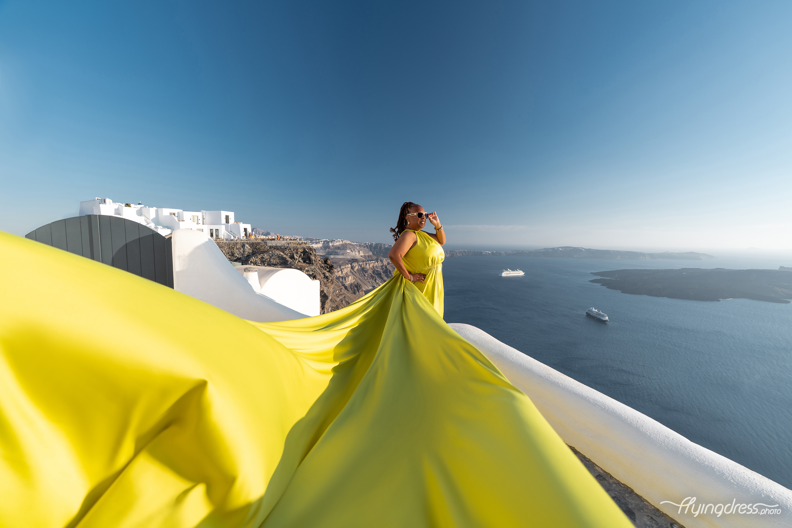 A vibrant neon yellow flying dress cascades over Santorini's white rooftops with the deep blue caldera stretching endlessly into the horizon.