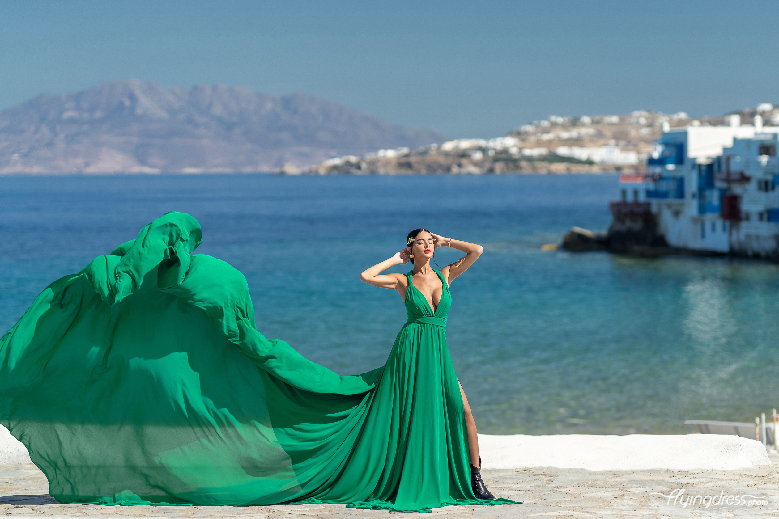 A woman in a flowing green dress poses confidently by the sea in Mykonos, Greece, with Little Venice and distant mountains in the background, as the wind lifts her dress dramatically.