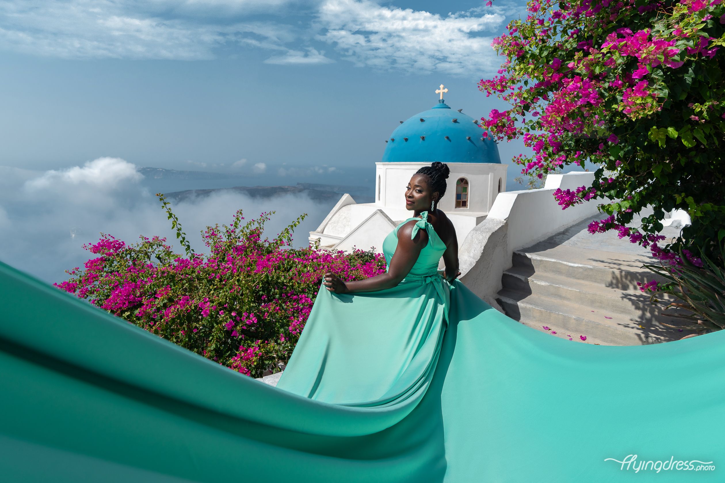 A stunning flying dress photoshoot in Santorini, featuring a woman in a flowing turquoise gown against iconic blue-domed churches and vibrant bougainvillea. Elegant, dreamy, and perfect for capturing the magic of the island.