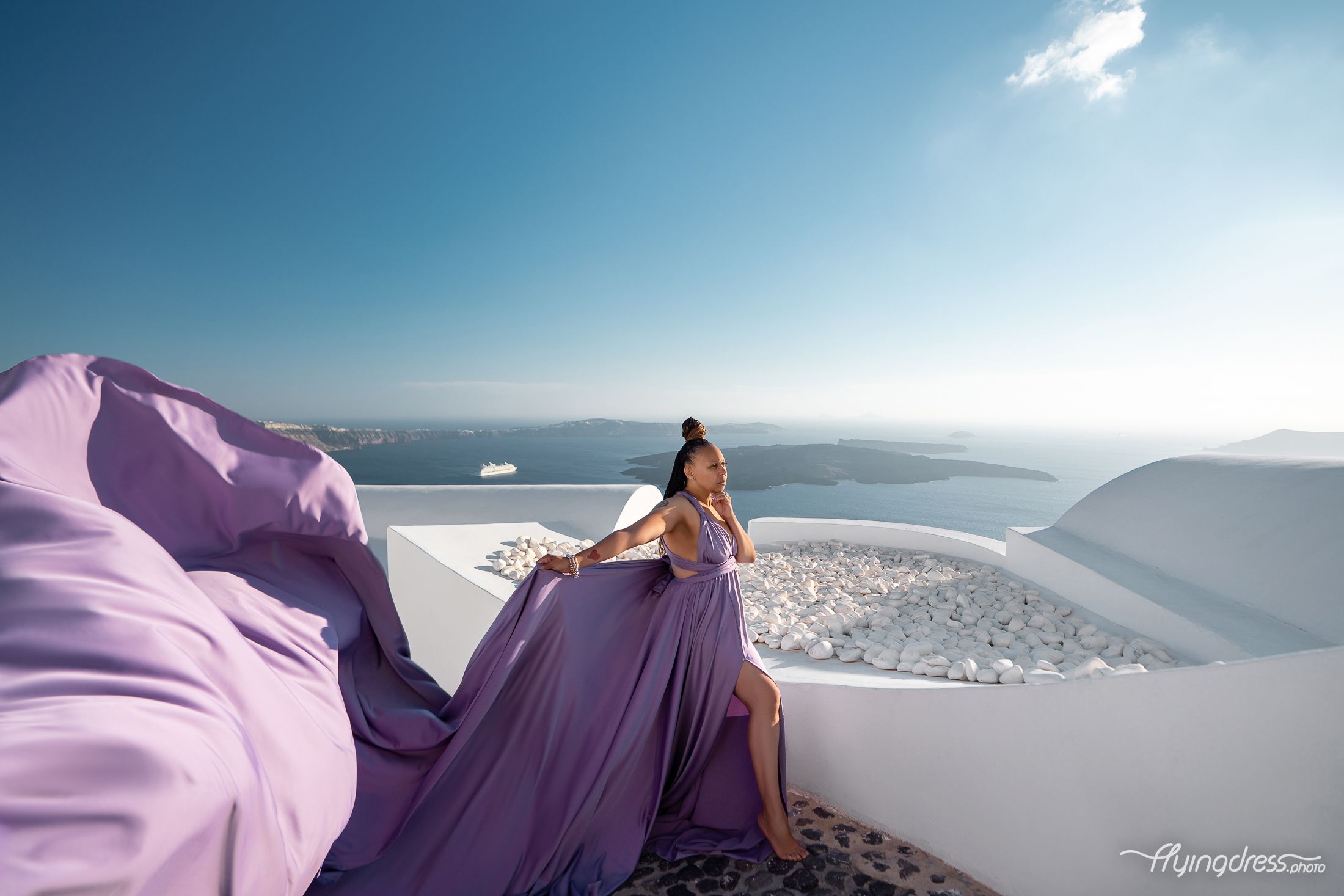 A dusty purple transformer dress billows over Santorini's sun-bleached terraces as the vast Aegean caldera melts into the horizon.
