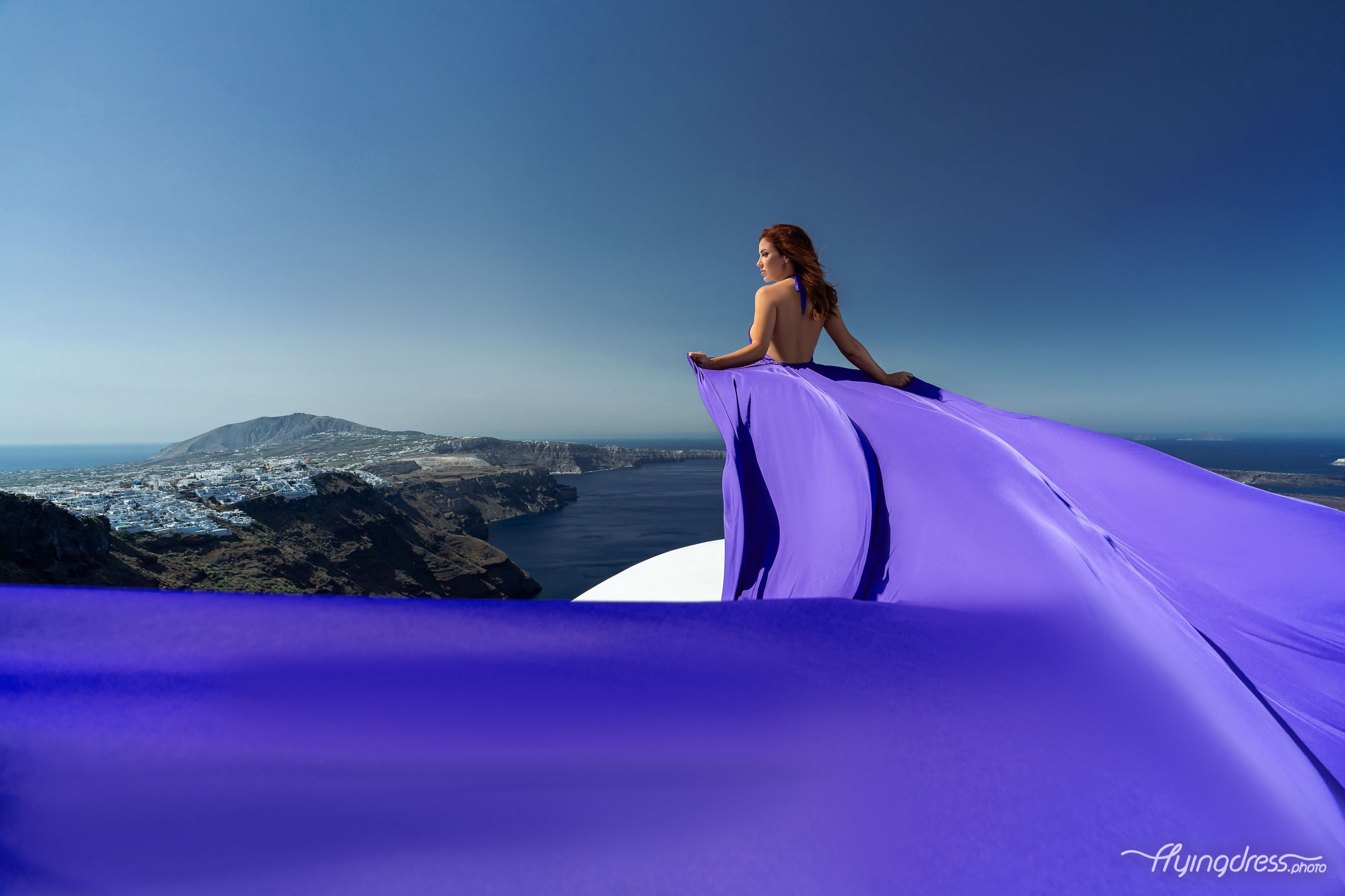 A woman in a flowing purple dress stands on a cliff overlooking a picturesque coastal landscape under a clear blue sky.