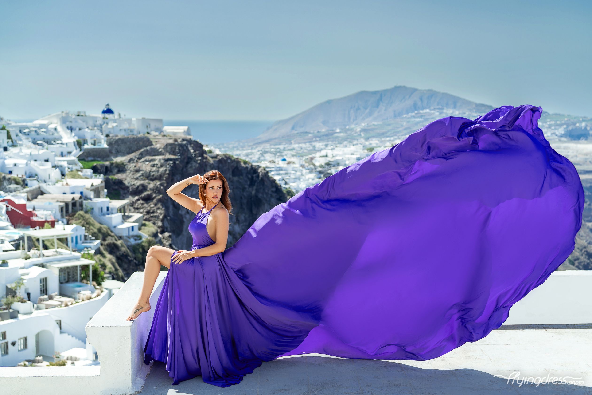 A woman in a stunning, flowing purple dress poses gracefully on a terrace with a scenic backdrop of white buildings and mountains by the sea.