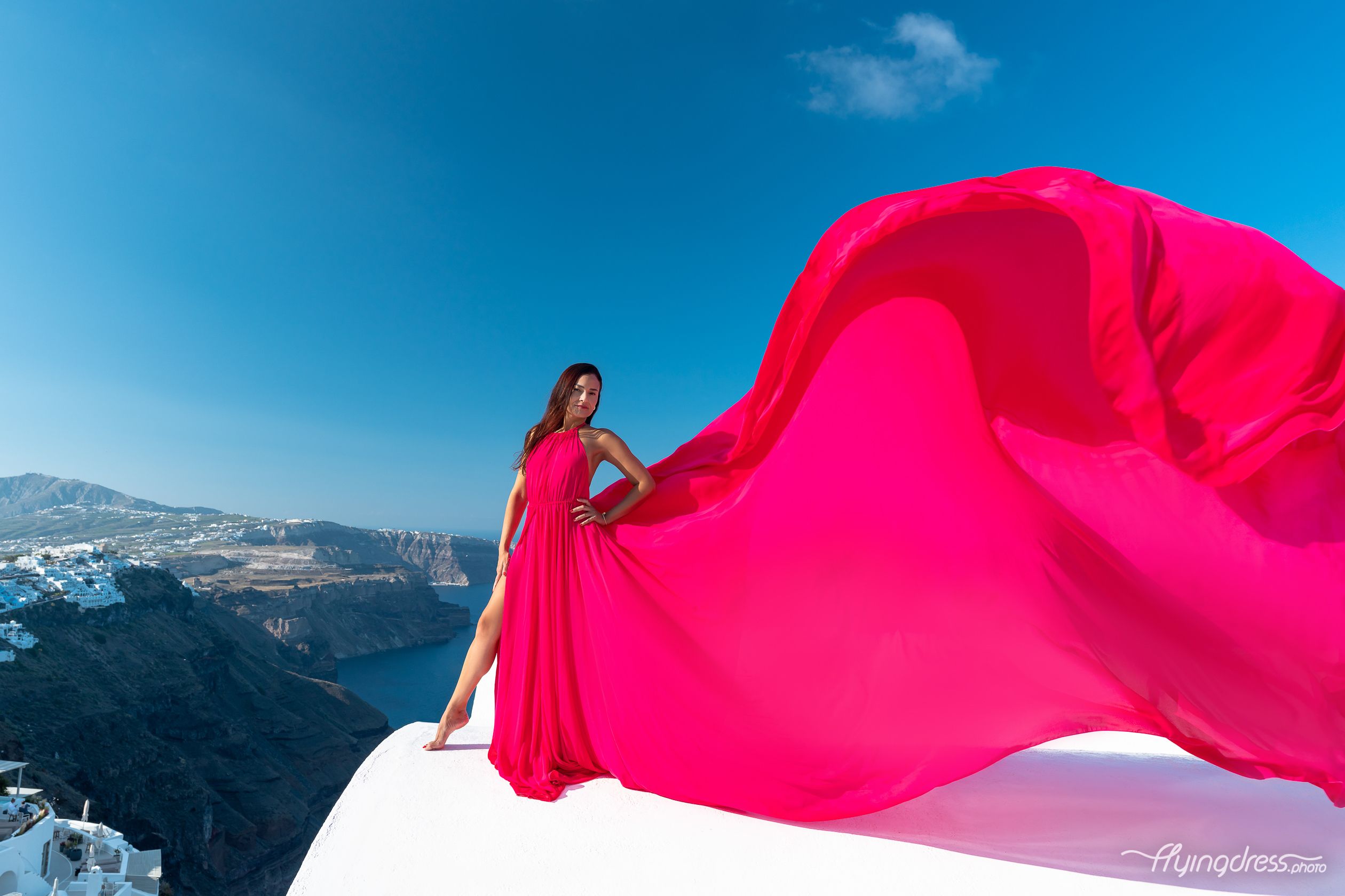 A lady in a fuchsia pink flying dress exudes a vibrant allure, adding a burst of color to the picturesque scenery of Santorini's landscapes.