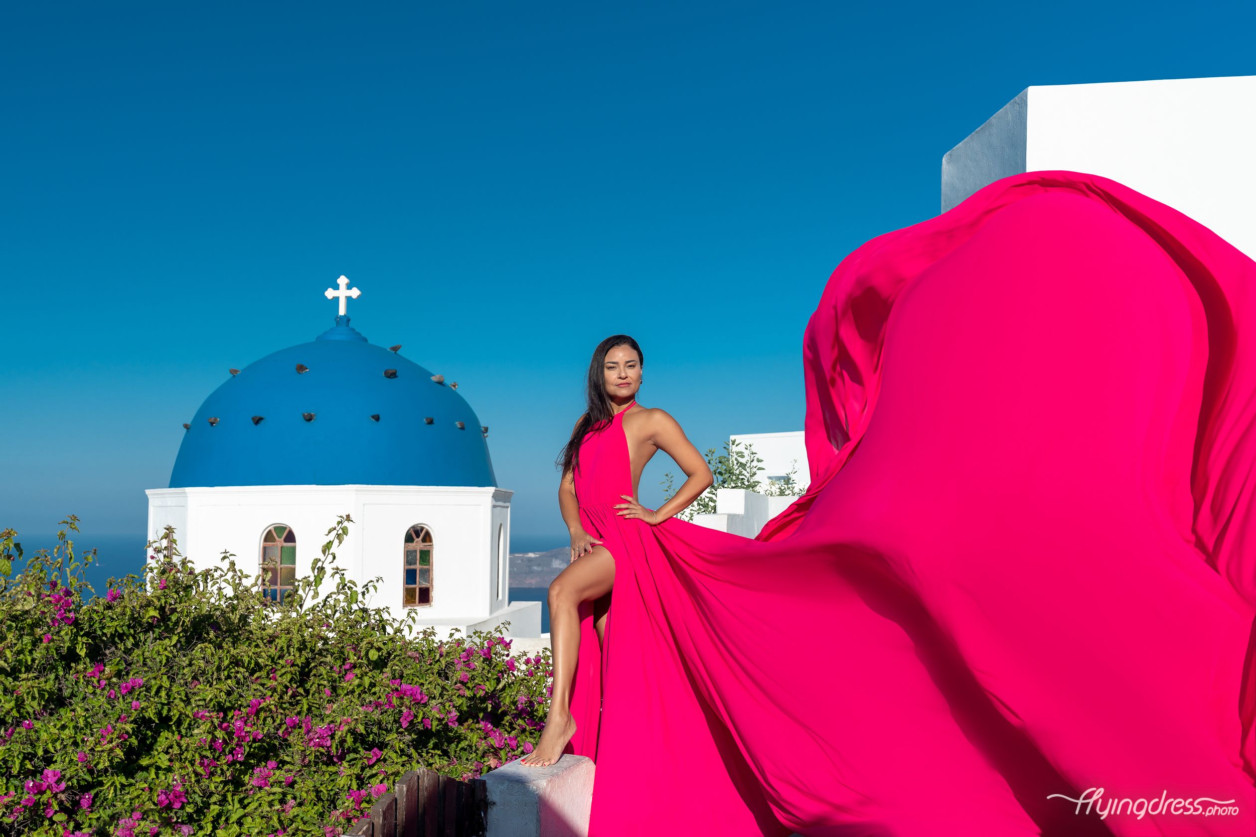 A vision of elegance and grace, the lady's fuchsia pink flying dress dances harmoniously with the wind, painting a striking picture against Santorini's backdrop.
