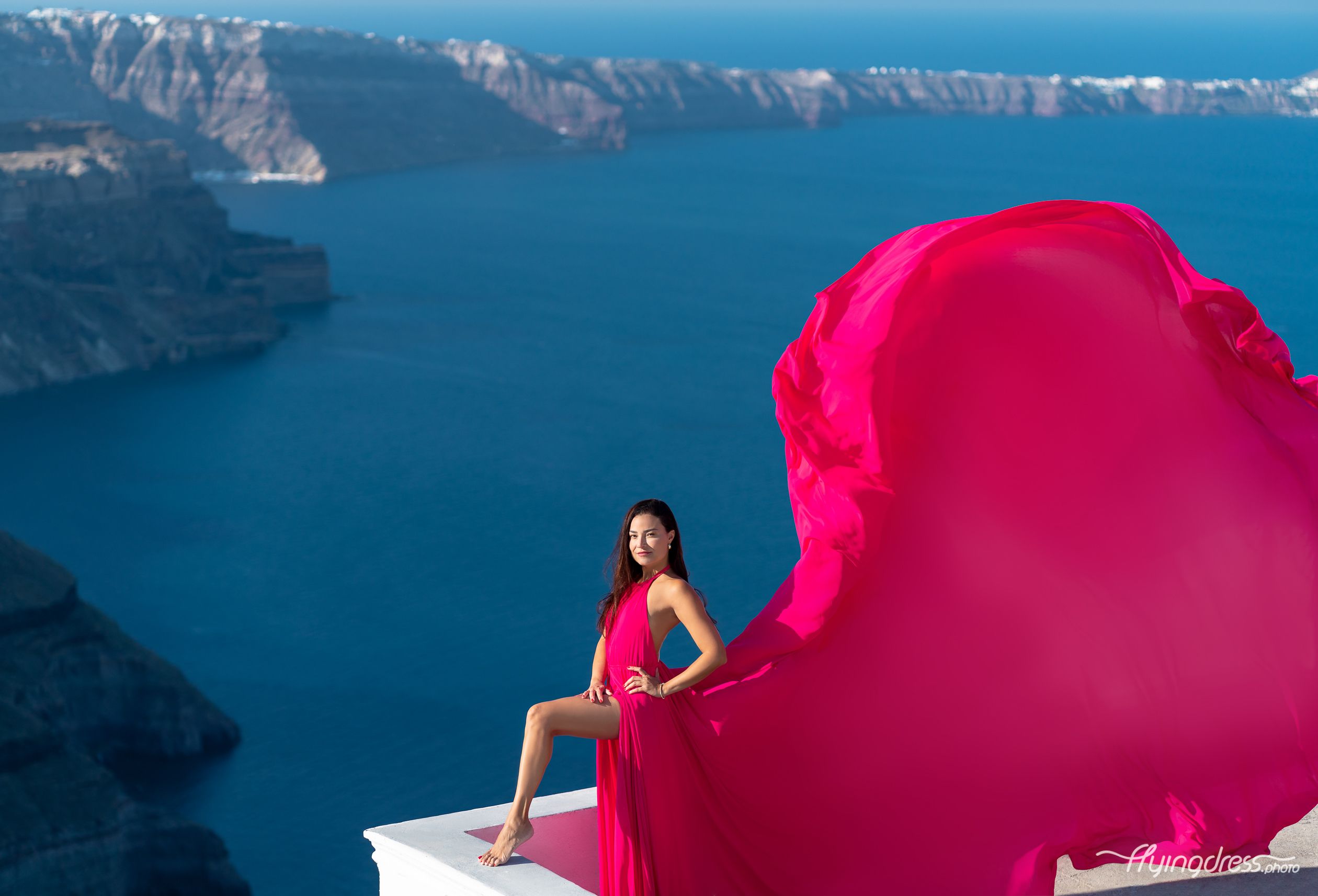 Gracefully adorned in a vibrant fuchsia pink flying dress, a lady becomes a vision of elegance and movement, her attire flowing with whimsical beauty against the backdrop of Santorini's breathtaking landscapes, capturing the essence of freedom and enchantment.
