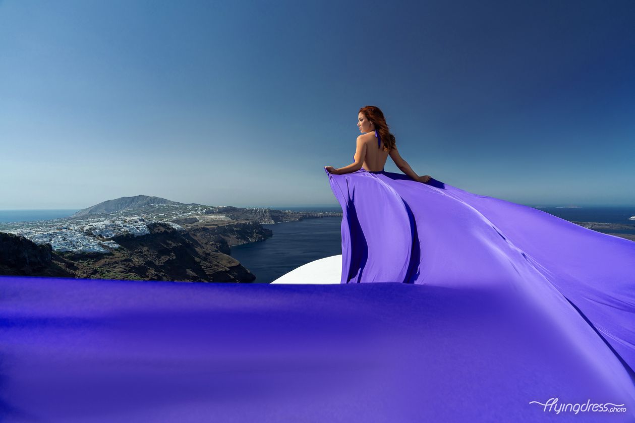 A woman in a flowing purple dress stands on a cliff overlooking a picturesque coastal landscape under a clear blue sky.