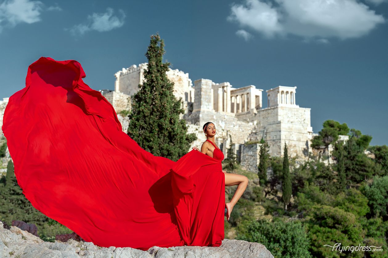 Standing against the backdrop of the Acropolis, a model in a striking red dress captivates in a brief but powerful moment, embodying elegance amidst Athens' timeless allure