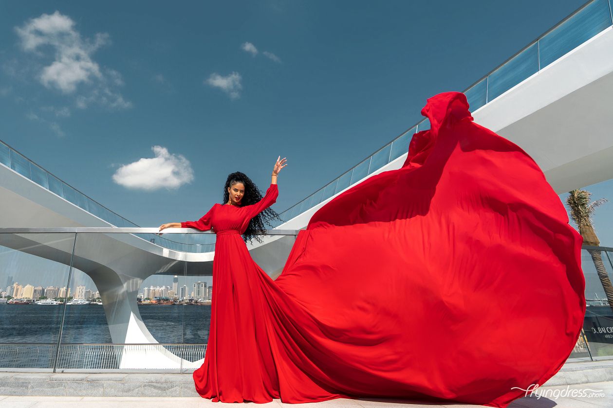 Young woman in a striking red dress gracefully leaps in Dubai Creek Harbor, embodying freedom and enchantment during a captivating photoshoot by the waterfront.
