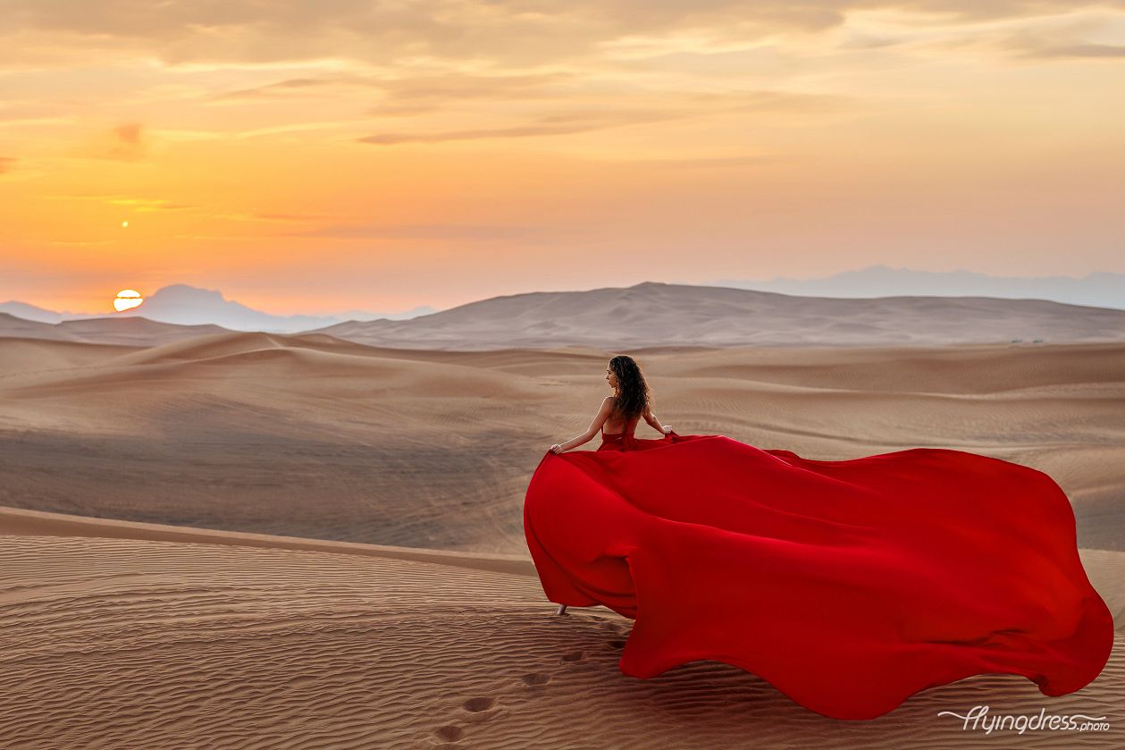 Captivating desert sunset photoshoot featuring a woman in a red flying dress, where the vibrant hues paint a mesmerizing backdrop to her graceful pose.
