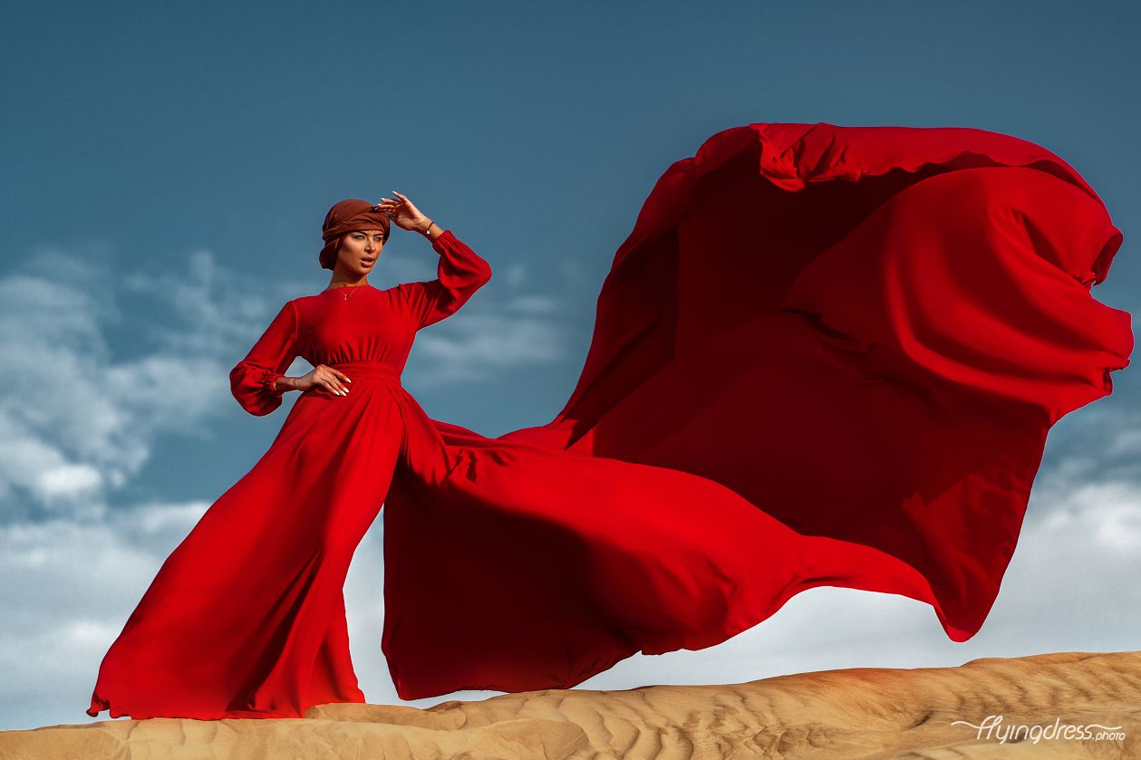Young woman in a stunning red dress gracefully leaps in Dubai's desert, embodying freedom and enchantment during a captivating photoshoot.