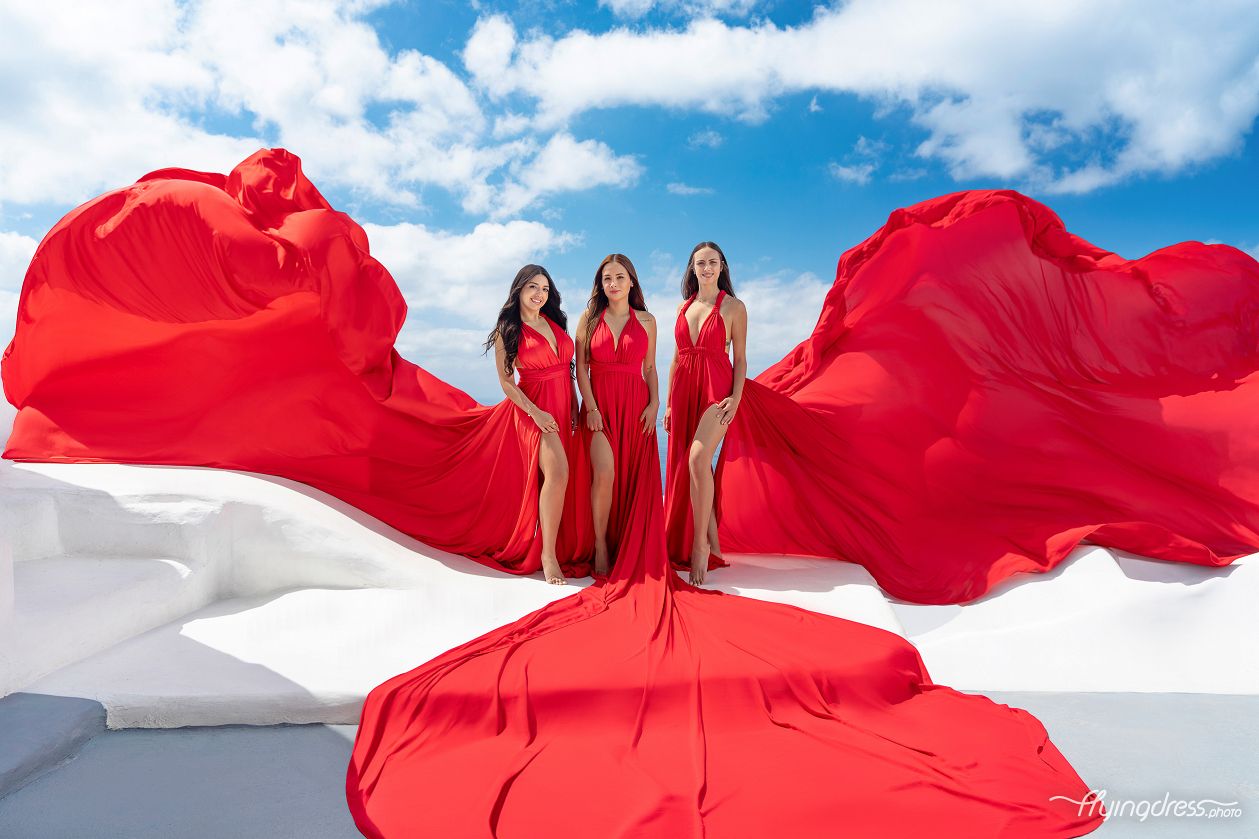Three women in flowing red gowns stand against a bright blue sky with scattered clouds