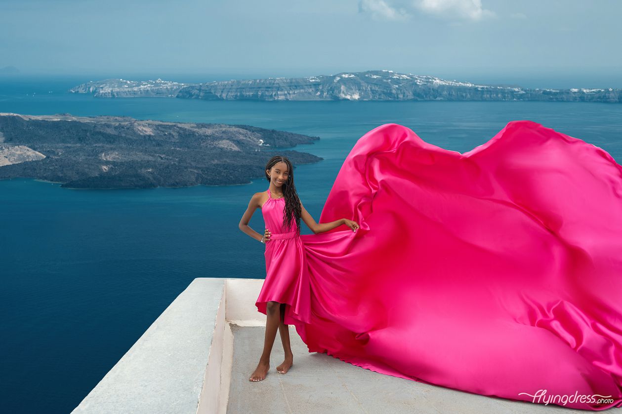 Girl in fuchsia satin flying dress standing in Santorini with a backdrop of the volcano.