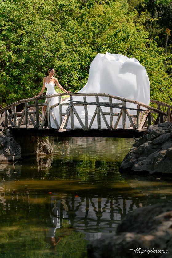 By the lake's edge, on a wooden bridge, a model in a flowing white dress exudes serene elegance, capturing timeless beauty in a National Garden photoshoot