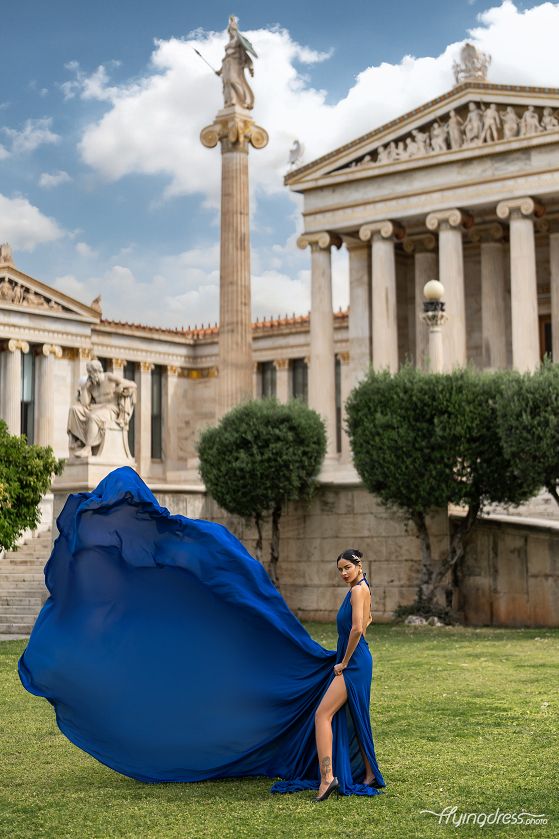 Adorning a radiant blue dress, a model poses amidst the historic splendor of Athens, infusing the scene with a captivating blend of modern fashion and timeless allure
