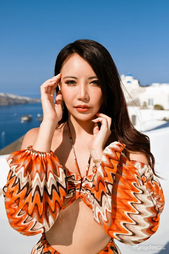 Against the backdrop of Oia's stunning vistas, an Asian girl in a vibrant, colorful dress radiates joy and charm, her presence adding an extra touch of vivacity and cultural richness to the picturesque streets of Santorini.