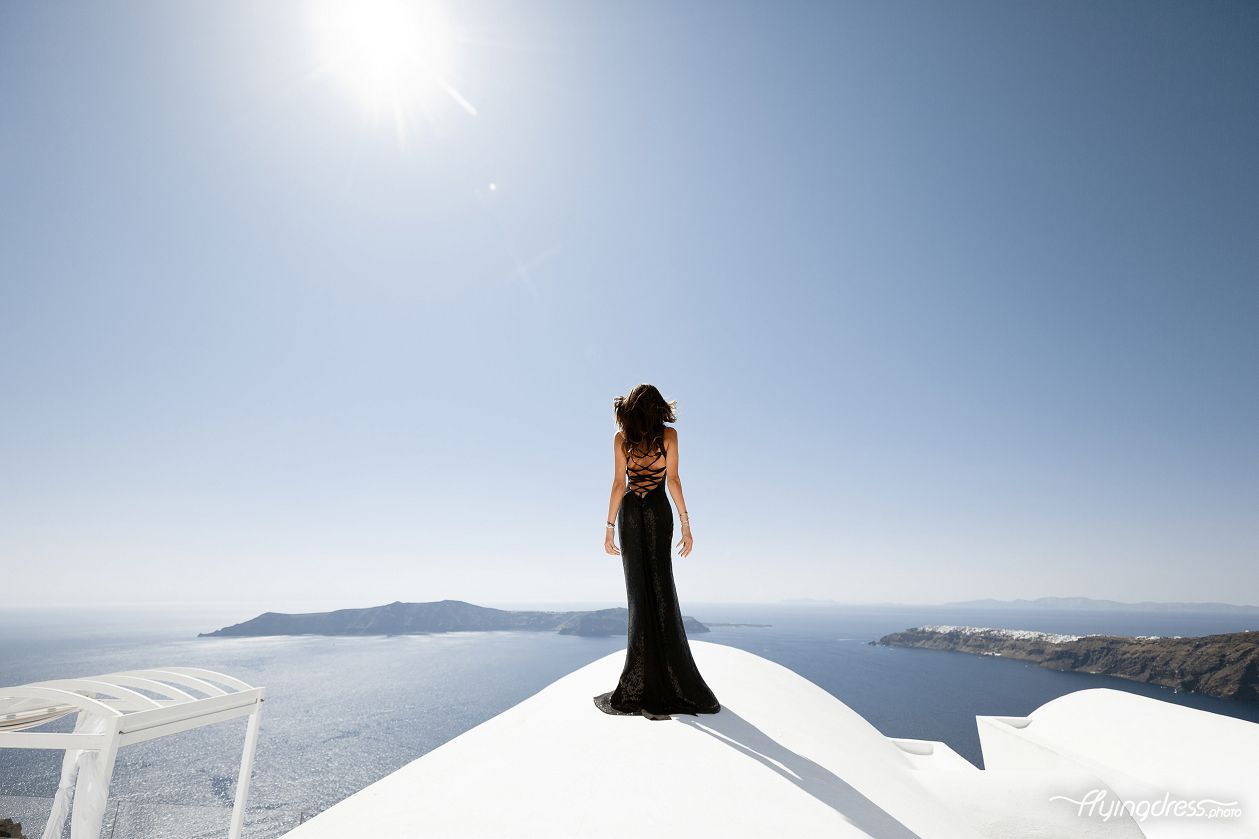 Against the backdrop of Santorini's breathtaking caldera view, a captivating girl in a flowing black dress stands poised, merging elegance with the awe-inspiring beauty of the island's panoramic vistas.