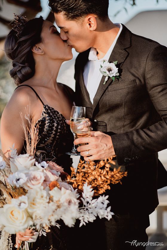 Locked in a passionate kiss, an Asian couple dressed in black attire celebrates their love amidst the scenic splendor of Santorini, their connection igniting a moment of pure romance and tenderness.
