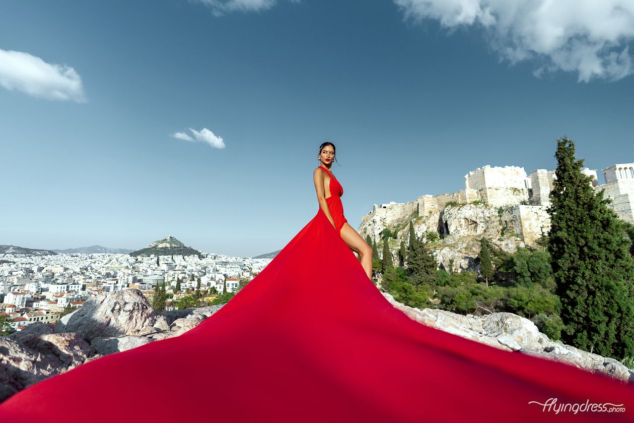 A striking silhouette: a model in a flowing red dress poses against the majestic backdrop of the Acropolis, with the bustling cityscape of Athens stretching out behind her during an enchanting photoshoot