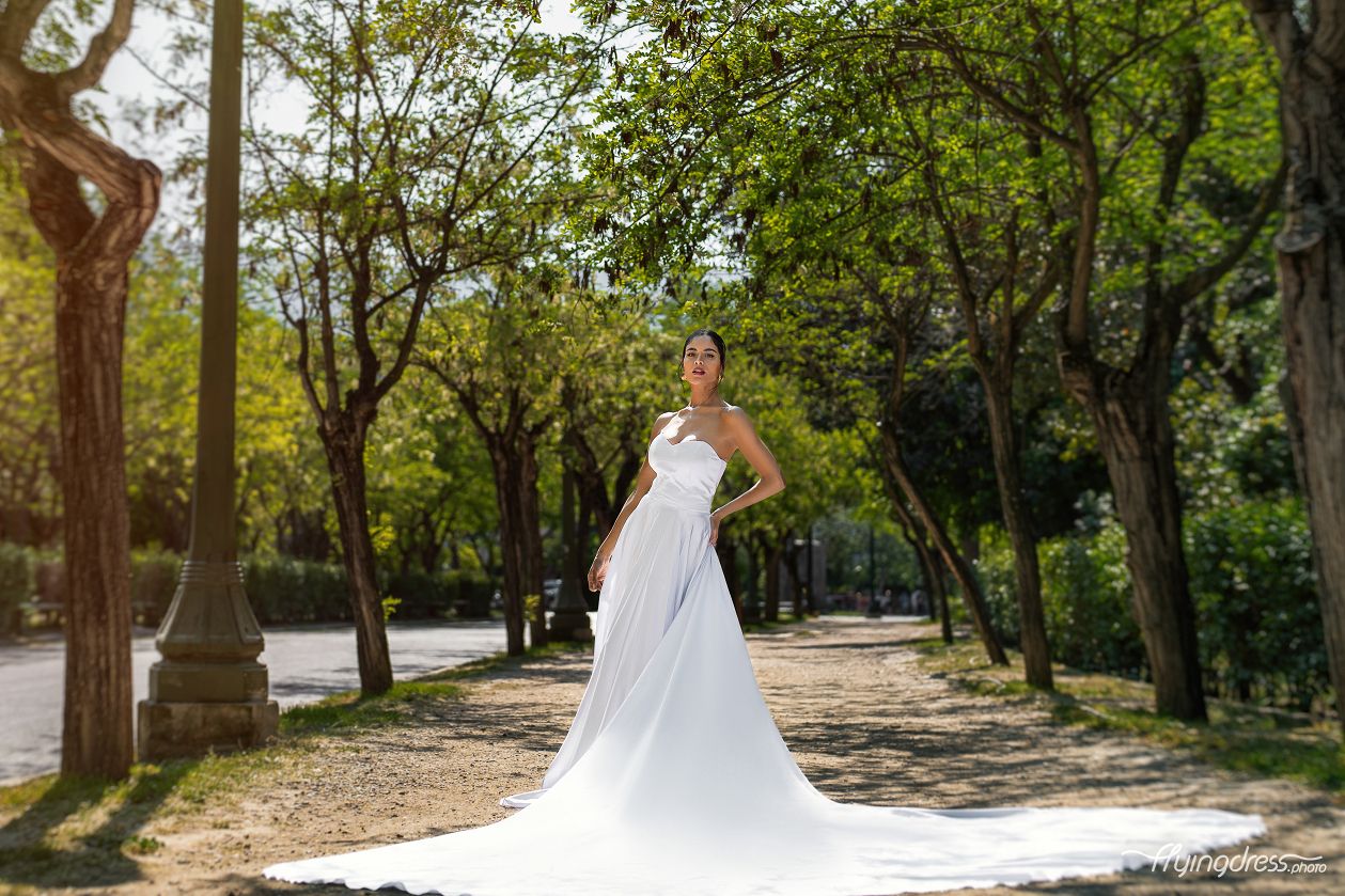 In Athens National Garden, a model dons a white corset flying dress, her ethereal presence adding a touch of enchantment to the serene surroundings during a captivating photoshoot