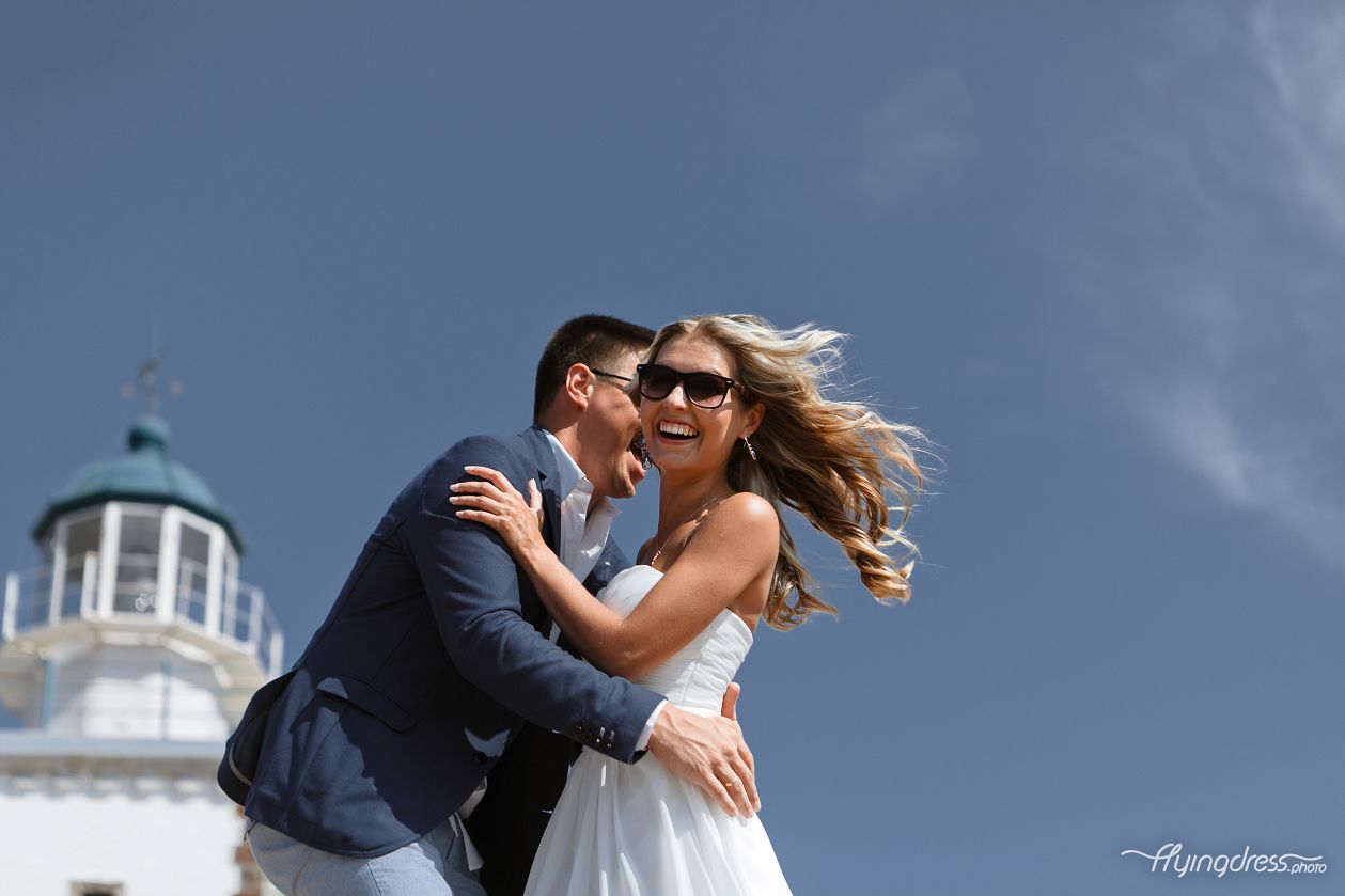 Amidst the romantic allure of Santorini's lighthouse, a wedding couple stands together, their smiles radiating happiness and their hearts entwined in a lifelong promise, as they create a cherished memory against the backdrop of this picturesque landmark.