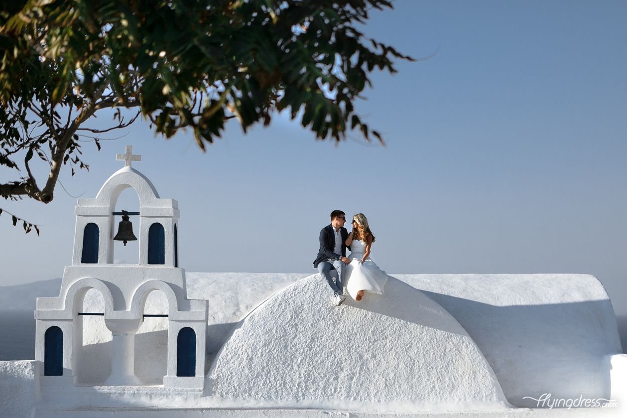 A couple sits intimately on a picturesque church in Santorini, their embrace blending the serenity of the sacred space with the depth of their love, creating a timeless moment of connection and devotion.