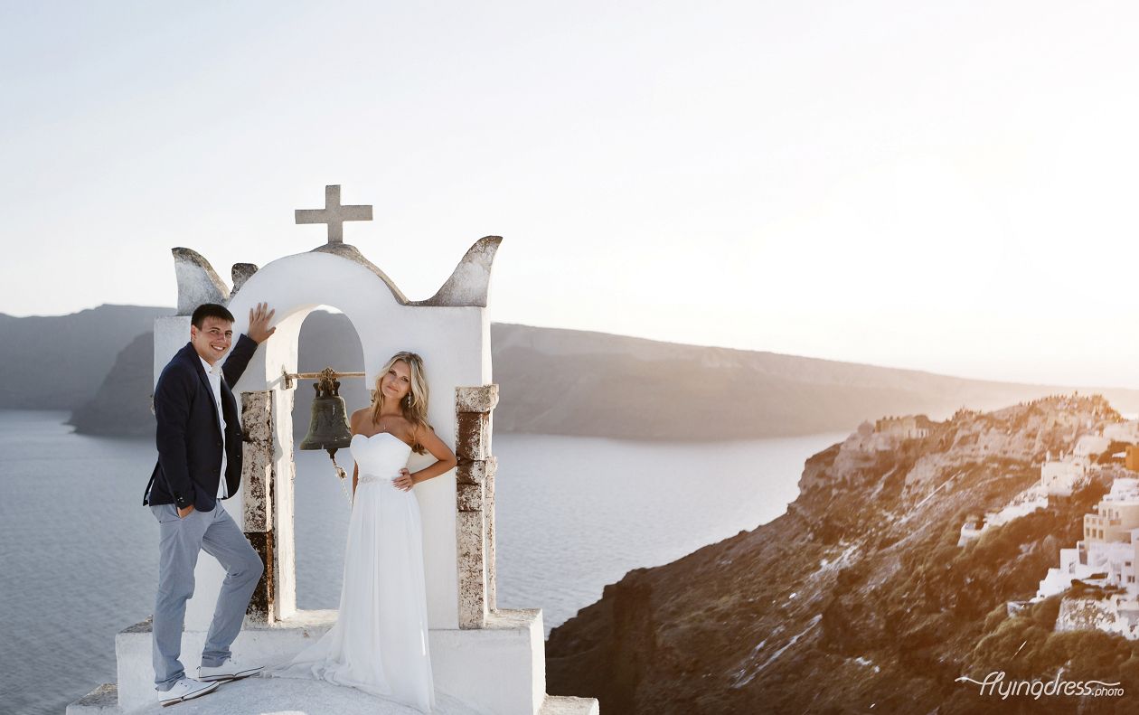 Against the backdrop of a stunning Santorini sunset, a couple stands close, their silhouettes embraced by the warm hues, embodying the timeless beauty of love and the breathtaking allure of this magical island.