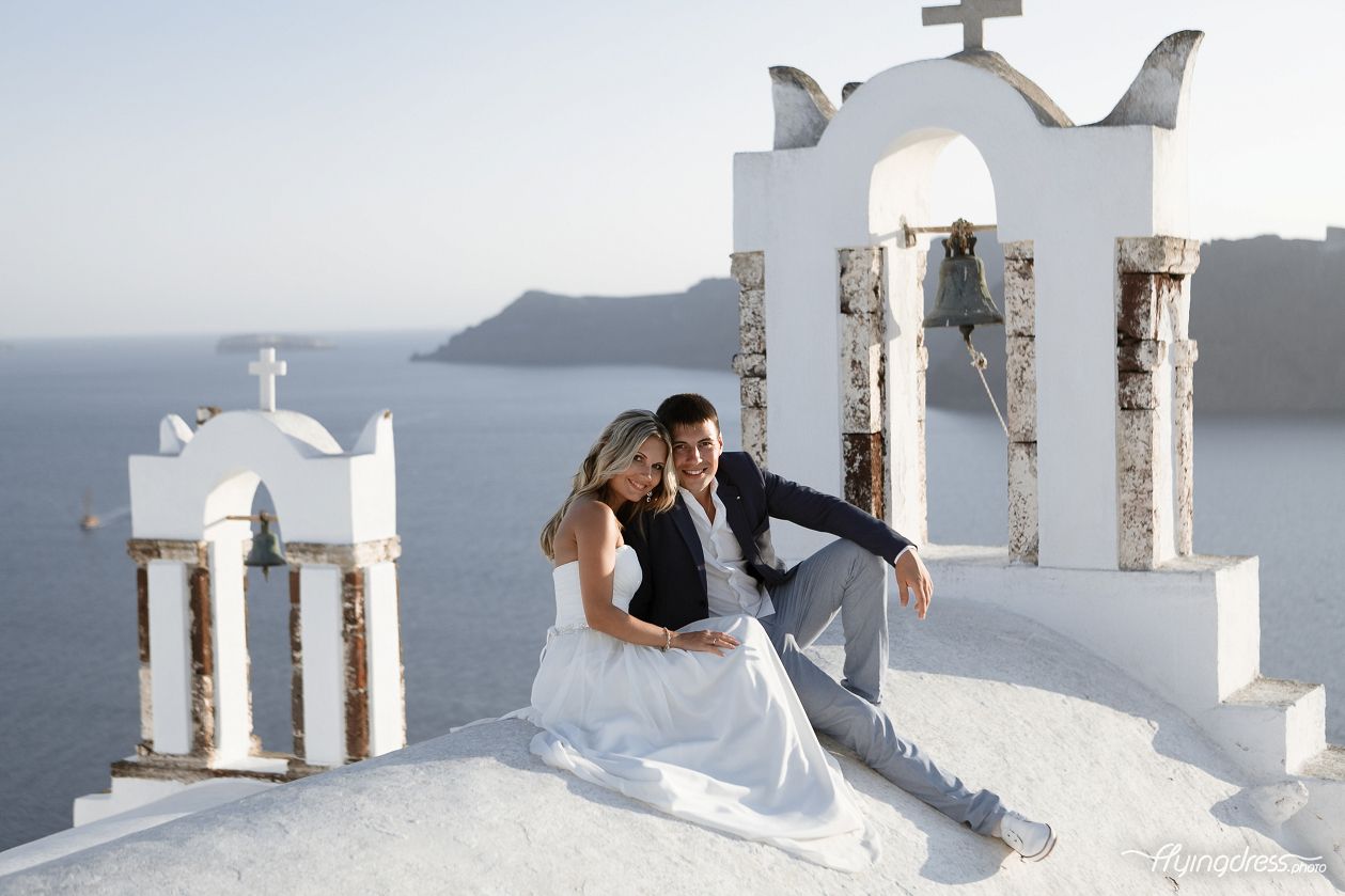 Seated together, a couple takes in the breathtaking view of Santorini, their love and contentment reflected in their smiles, as they find solace and joy in each other's company amidst the island's captivating beauty.