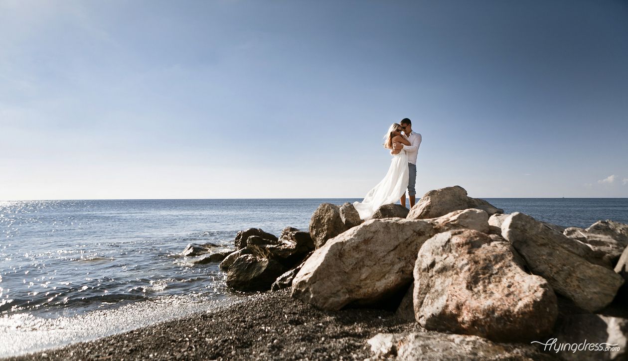 Standing on a rock at Santorini's black sandy beach, a couple embraces the rugged beauty of the volcanic landscape, their love and adventurous spirit amplified against the dramatic contrast of the dark sands and the shimmering sea.