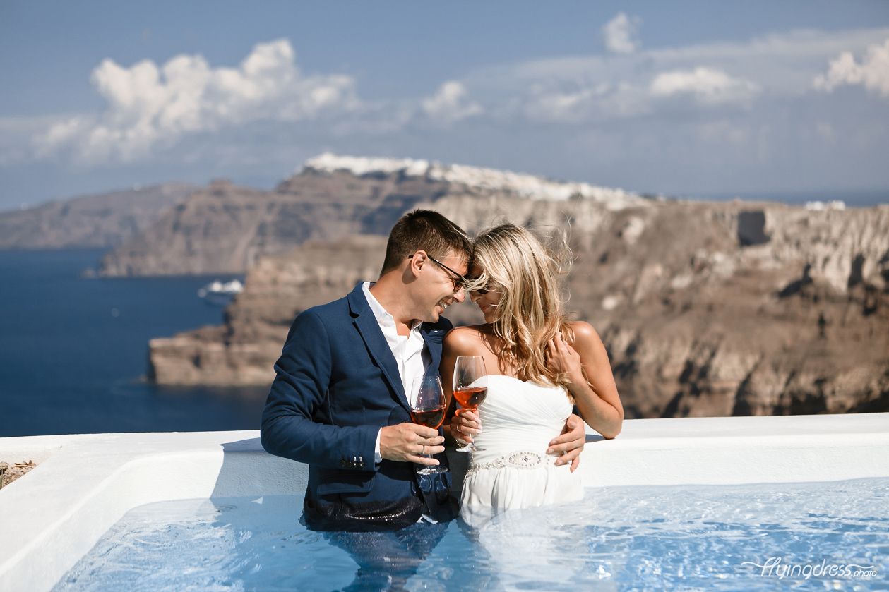 Immersed in the refreshing pool waters, a couple basks in the panoramic beauty of Santorini's caldera view, their laughter echoing through the air as they create cherished memories in this idyllic oasis.