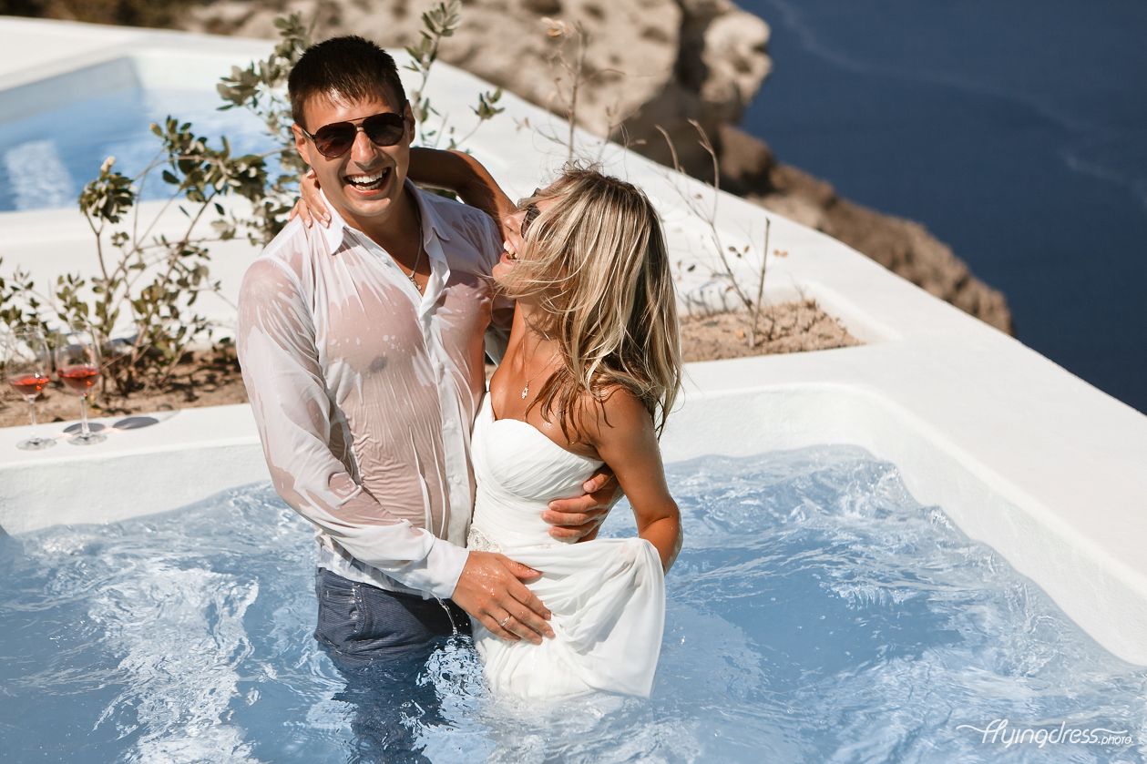 With their clothes drenched in water, a couple gleefully enjoys a playful moment in the pool, their laughter echoing through the air, embracing the spontaneity and exhilaration of the experience in Santorini's idyllic setting.