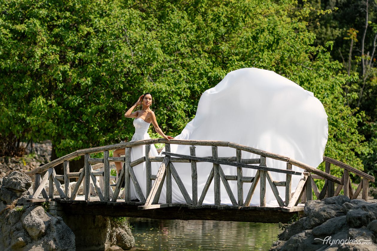 At the tranquil lakeside, upon a wooden bridge, a model poses gracefully in a flowing white dress, exuding serenity and charm amidst the natural beauty of the National Garden in a captivating photoshoot