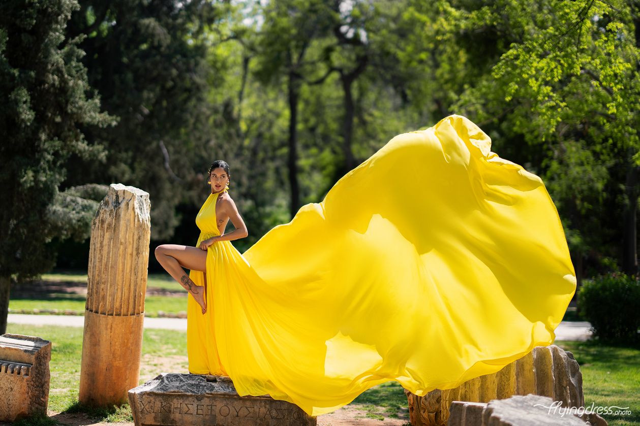 Amidst the verdant beauty of the Garden of Athens, a model graces the scenery in a flowing yellow dress, embodying elegance and vibrancy in a captivating photoshoot