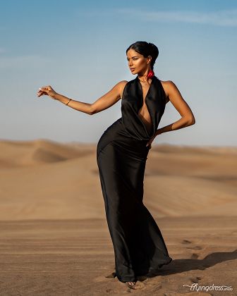 A woman in an elegant black dress strikes a confident pose in the vast, golden sand dunes of the Dubai desert, her look exuding poise and sophistication against the serene backdrop.