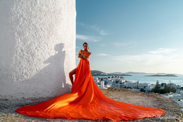 A woman in a vibrant orange gown poses elegantly against the backdrop of a traditional white-washed building in Mykonos, with her dress flowing dramatically and the picturesque Aegean Sea and island landscape in the distance.