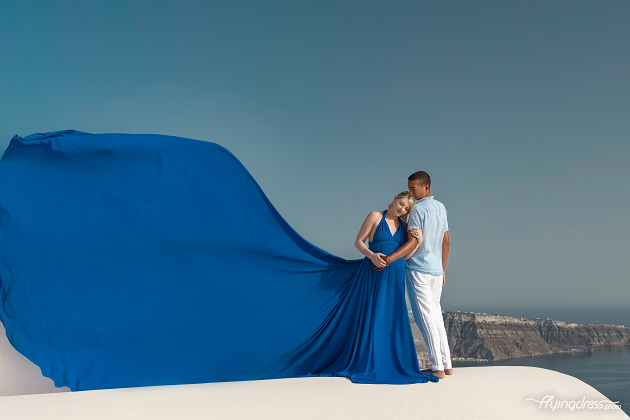 A couple poses for a serene maternity photoshoot in Santorini, Greece, with the woman wearing a flowing blue dress that billows dramatically in the wind.