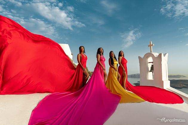Four women in vibrant flowing dresses stand on a white rooftop with a cross, overlooking a scenic coastal view under a bright blue sky.