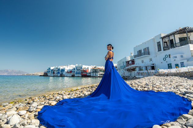 A woman in a flowing blue dress stands gracefully on a rocky beach, with the picturesque waterfront buildings of Mykonos' Little Venice in the background.