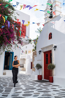 A woman in a sleek black outfit walks through a colorful alley in Mykonos, surrounded by festive flags and vibrant whitewashed buildings.