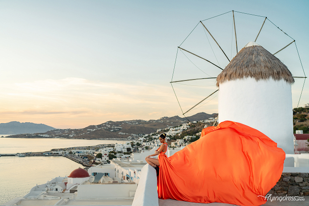 A woman in a flowing orange gown poses gracefully in front of a traditional windmill in Mykonos, with the soft hues of the sunset illuminating the white-washed buildings and serene Aegean Sea in the background.